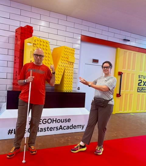 Victor, a man with a white cane, and Alina, a woman with glasses, stand on a red carpet in front of a giant